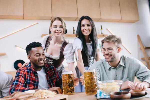 Two beautiful waitresses in traditional german costumes serving beer for multicultural friends in pub — Stock Photo