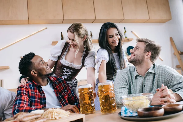 Two cheerful waitresses in traditional german costumes serving beer for multicultural friends in pub — Stock Photo