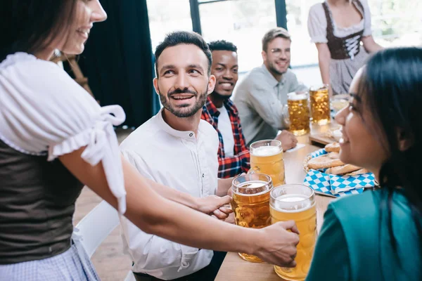 Cropped view of waitresses in traditional german costumes serving beer for multicultural friends in pub — Stock Photo