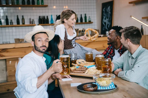 Homem bonito na palha segurando copo de cerveja e olhando para a câmera enquanto celebra octoberfest com amigos multiculturais — Fotografia de Stock