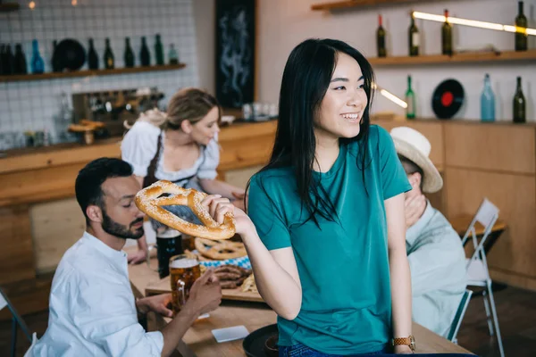 Selective focus of asian girl holding pretzel while celebrating octoberfest with multicultural friends — Stock Photo