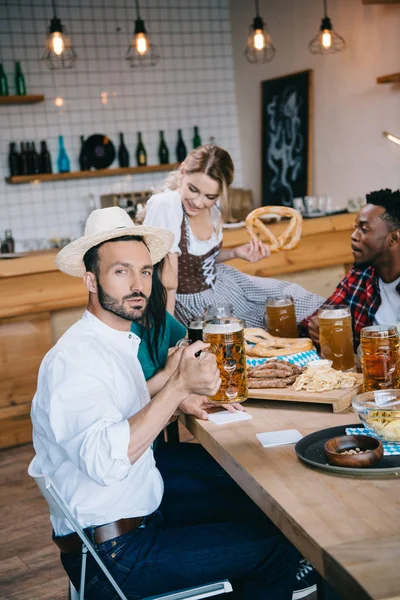 Young man in straw holding glass of beer and looking at camera while celebrating octoberfest with multicultural friends — Stock Photo