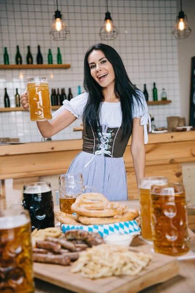 Hermosa joven camarera en traje alemán tradicional sosteniendo taza de cerveza ligera y sonriendo a la cámara - foto de stock