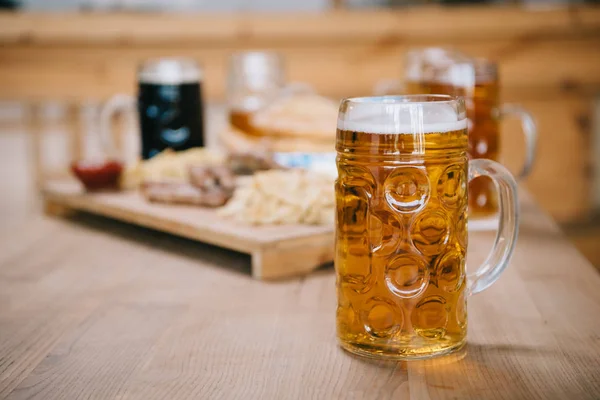 Selektiver Fokus des Bechers mit Lagerbier auf Holztisch in der Nähe des Tabletts mit Snacks in der Kneipe — Stockfoto