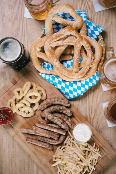Vista superior de salchichas fritas, aros de cebolla, papas fritas, pretzels y tazas con cerveza en mesa de madera en pub - foto de stock