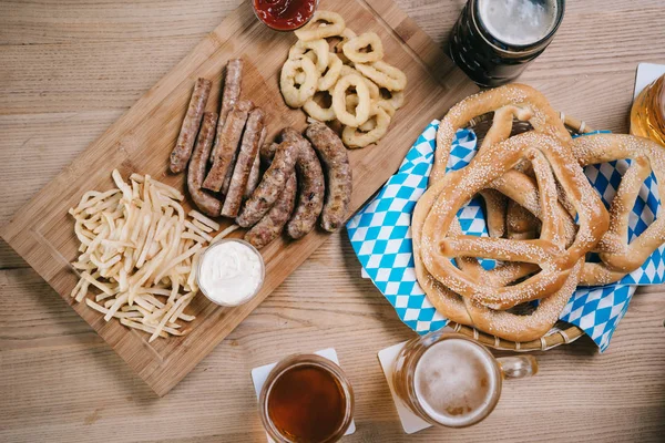 Top view of mugs with light and dark beer, fried sausages, onion rings, french fries and pretzels on wooden table in pub — Stock Photo