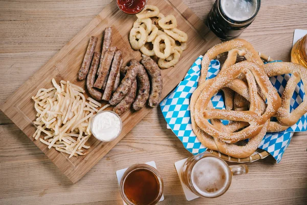 Vista superior de canecas com cerveja clara e escura, salsichas fritas, anéis de cebola, batatas fritas e pretzels na mesa de madeira no pub — Fotografia de Stock