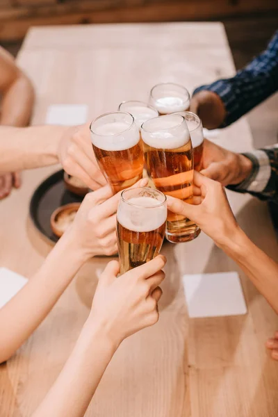 Vista parcial de amigos multiculturales tintineo vasos con cerveza lager en el pub - foto de stock