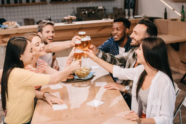 Cheerful multicultural friends clinking glasses with beer while celebrating octoberfest in pub — Stock Photo