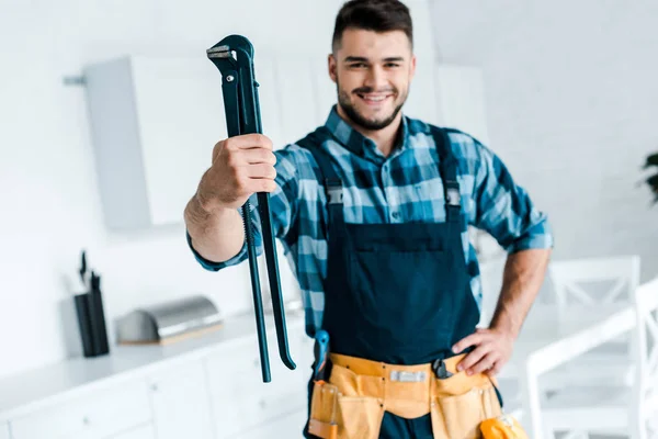 Selective focus happy handyman in uniform standing with hand on hip and holding wire cutters — Stock Photo