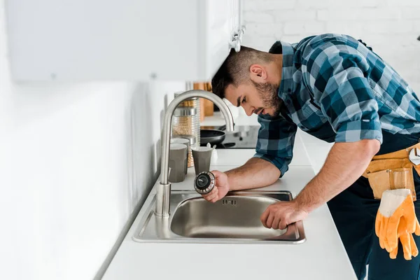 Selective focus of handsome bearded repairman working near sink in kitchen — Stock Photo