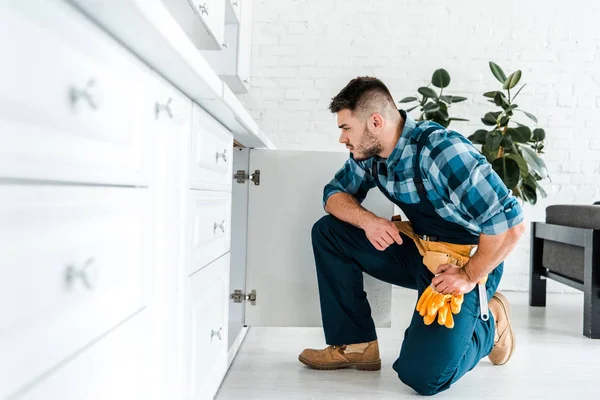 Selective focus of handsome handyman with tool belt sitting near kitchen cabinet — Stock Photo