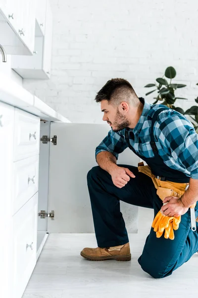 Vista lateral do handyman bonito com cinto de ferramentas sentado perto do armário da cozinha — Fotografia de Stock
