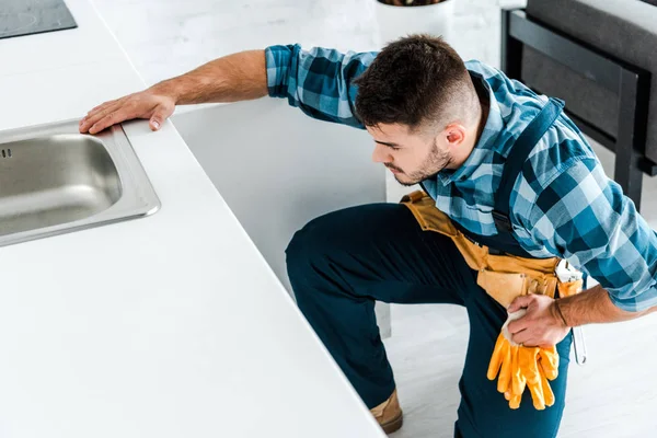 Bearded repairman with tool belt sitting near kitchen cabinet — Stock Photo