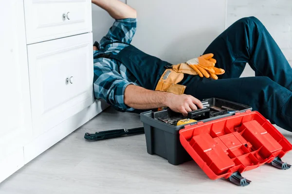 Vue recadrée du réparateur travaillant dans la cuisine près de la boîte à outils tout en étant couché sur le sol — Photo de stock