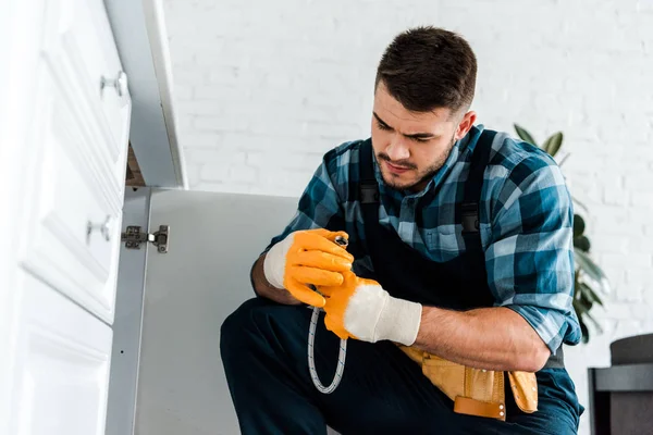 Selective focus of repairman looking at metal hose near kitchen cabinet — Stock Photo