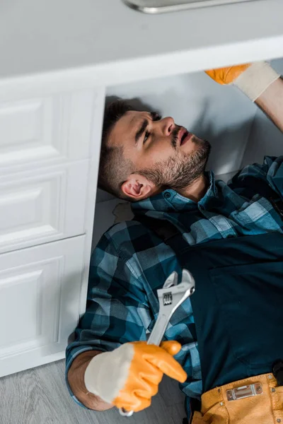 Selektiver Fokus des gutaussehenden Reparateurs mit verstellbarem Schraubenschlüssel während der Arbeit in der Küche — Stockfoto