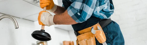 Panoramic shot of handyman with tool belt holding plunger in kitchen — Stock Photo