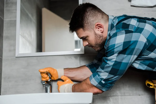Handsome bearded repairman standing near sink and touching faucet in bathroom — Stock Photo