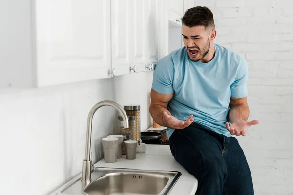 Расстроенный мужчина жестом и криком возле крана на кухне — стоковое фото