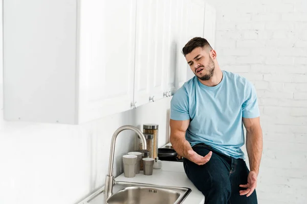 Selective focus of upset man looking at faucet and sink in kitchen — Stock Photo