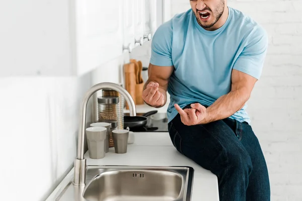 Vue recadrée de l'homme contrarié montrant les doigts du milieu et criant près du robinet dans la cuisine — Photo de stock
