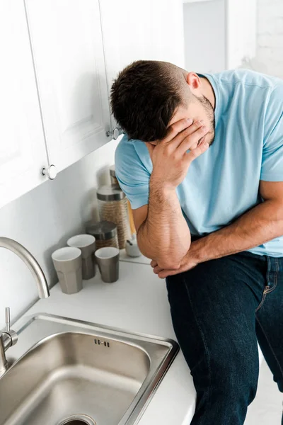 Homme frustré couvrant le visage tout en étant assis près du robinet dans la cuisine — Photo de stock