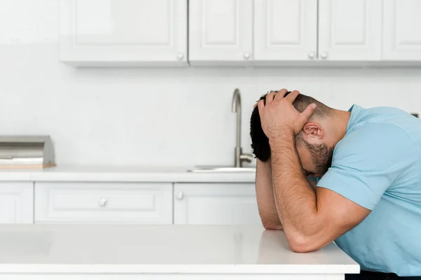 Разочарованный мужчина касается головы, сидя рядом со столом на кухне — стоковое фото