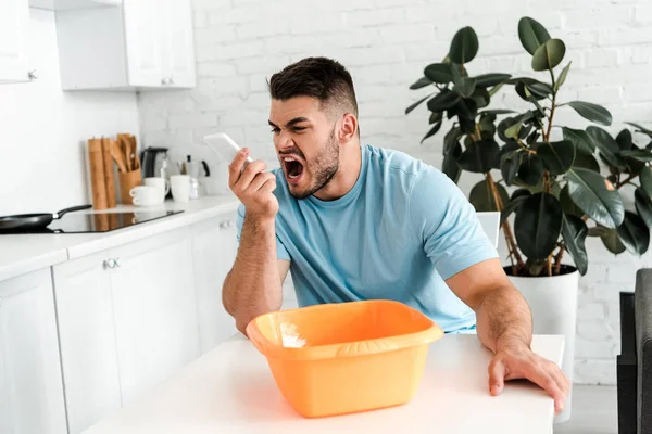 Fuoco selettivo di uomo barbuto sconvolto urlando allo smartphone vicino alla ciotola di plastica lavaggio — Foto stock