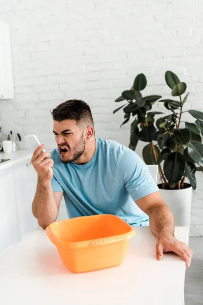 Selective focus of upset bearded man screaming while holding smartphone near water pouring into plastic wash bowl — Stock Photo