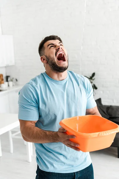 Messa a fuoco selettiva di uomo sconvolto urlando mentre tiene ciotola di plastica lavaggio vicino versando acqua — Foto stock