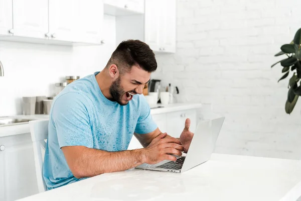 Hombre emocional con los ojos cerrados gritando cerca del ordenador portátil en la cocina - foto de stock