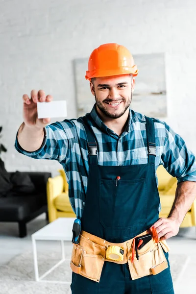 Trabajador feliz sosteniendo la tarjeta en blanco y de pie con la mano en la cadera - foto de stock