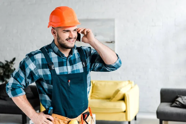 Cheerful worker in safety helmet talking on smartphone while standing with hand on hip — Stock Photo