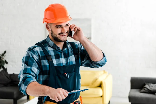 Fröhlicher Arbeiter im Schutzhelm hält Klemmbrett, während er mit dem Smartphone spricht — Stockfoto