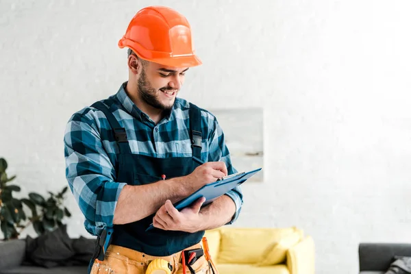 Cheerful bearded worker in safety helmet writing while holding clipboard — Stock Photo