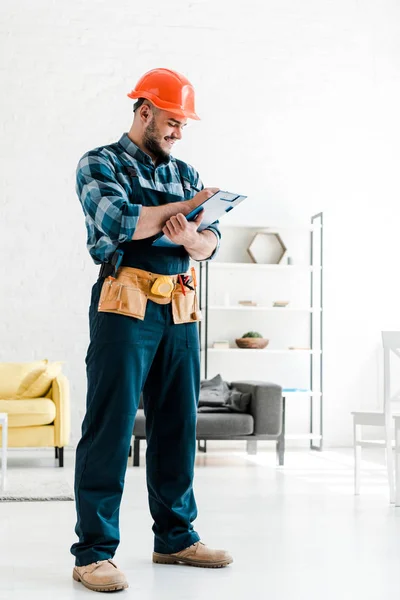 Happy bearded worker in safety helmet writing while holding clipboard in living room — Stock Photo