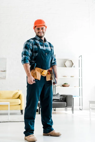 Glücklicher bärtiger Arbeiter mit Schutzhelm hält Klemmbrett im Wohnzimmer — Stockfoto