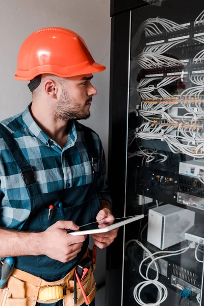 Handsome bearded workman holding digital tablet and looking at wires and cables — Stock Photo