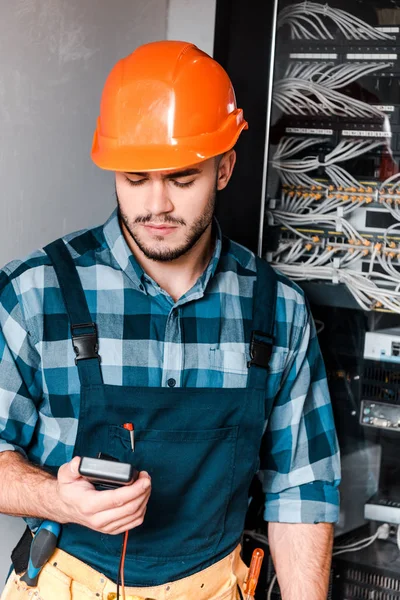 Handsome bearded technician holding digital meter near wires and cables — Stock Photo