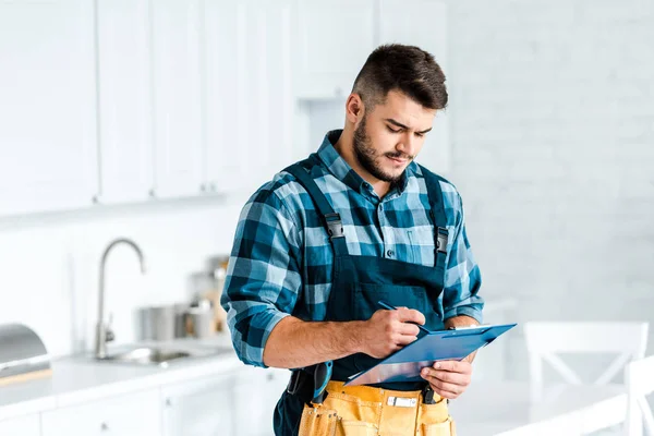 Bonito barbudo trabalhador escrevendo enquanto segurando prancheta na cozinha — Fotografia de Stock