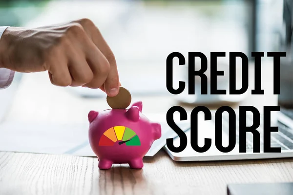Vista recortada de hombre de negocios poner moneda en alcancía rosa cerca de letras puntuación de crédito y portátil - foto de stock