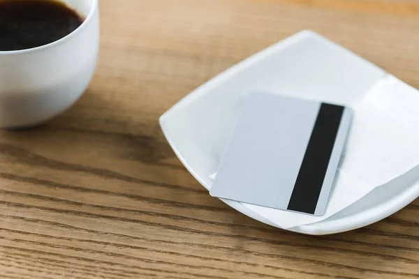 Credit card in white plate with receipt near cup with coffee on wooden table — Stock Photo