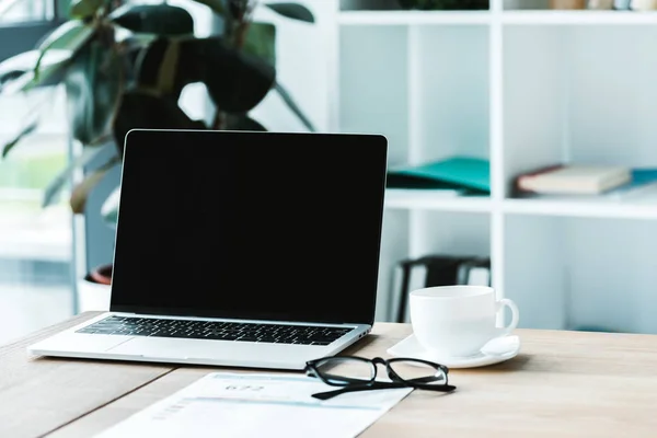 Tazza di caffè vicino al computer portatile con schermo bianco e bicchieri sul tavolo — Foto stock