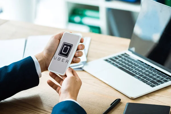 KYIV, UKRAINE - JULY 8, 2019: cropped view of man holding smartphone with uber app near laptop and pen on table — Stock Photo