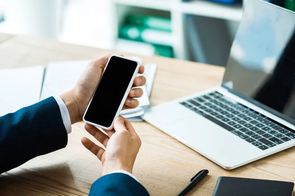 Ausgeschnittene Ansicht eines Geschäftsmannes, der ein Smartphone mit leerem Bildschirm in der Nähe des Laptops auf dem Tisch hält — Stockfoto