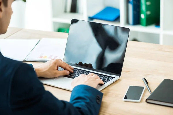 Selective focus of man typing on laptop near smartphone with blank screen on table — Stock Photo