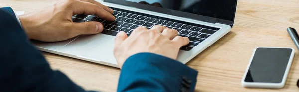 Panoramic shot of businessman typing on laptop near smartphone with blank screen — Stock Photo