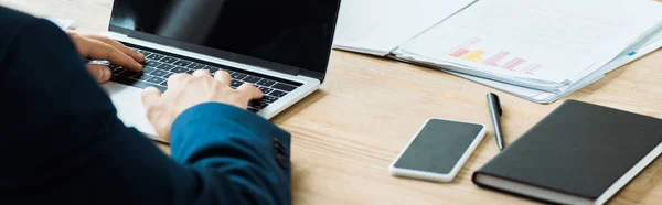 Plano panorámico del hombre escribiendo en el ordenador portátil cerca de teléfono inteligente con pantalla en blanco - foto de stock