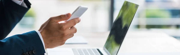 Panoramic shot of man holding smartphone near laptop — Stock Photo